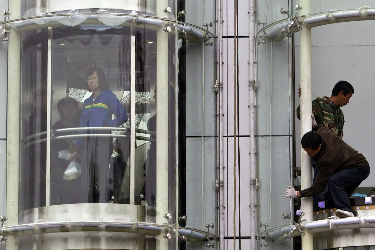 FILE - In this Oct. 21, 2010 file photo, workers, right, prepare to install a piece of glass wall covering an elevator tunnel while a woman, left, looks down from a descending elevator in Beijing, China. Otis elevator, which has staked much on increasing urbanization and soaring skyscrapers in cities across China, is struggling with declining revenue in 2015 as China’s economy sours. For Otis, based in Farmington, Conn., rising competition, particularly for the lucrative repair and maintenance business, also is undermining its Chinese market share.