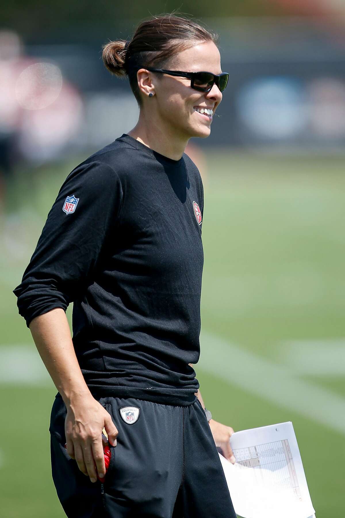San Francisco 49ers assistant coach Katie Sowers smiles as she watches players run through drills during practice at the San Francisco 49ers training facility on Wednesday, August 23, 2017, in Santa Clara California. Sowers, 31, made more history, becoming the NFL's first openly gay coach. San Francisco 49ers officially hired Katie Sowers for the 2017 season last week, making her the team's first female assistant coach.