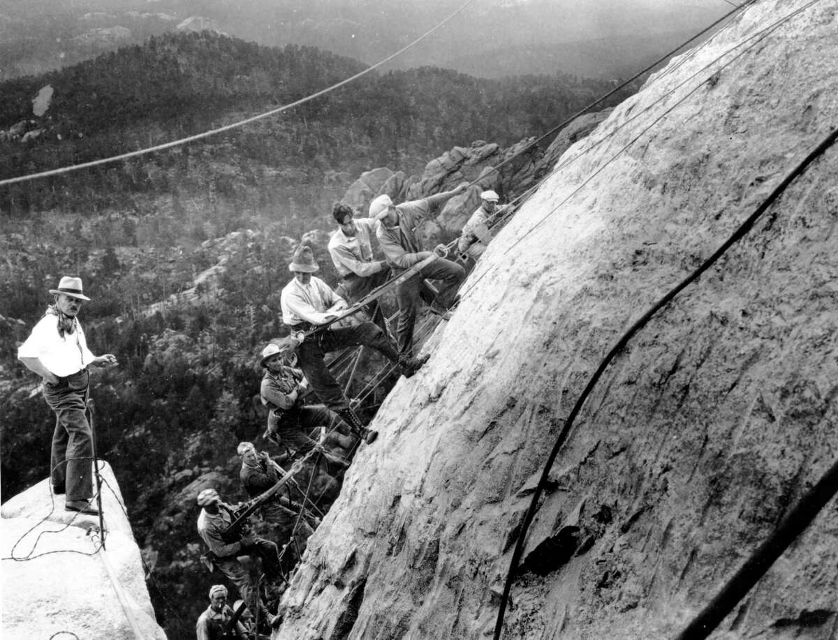 In this July, 22, 1929 photo, sculptor Gutzon Borglum, at left, directs drillers suspended by cables from the top of the mountain as they work on the head of President George Washington at the Mount Rushmore Memorial in the Black Hills area near Keystone, S.D. In October 2016, the memorial that through the years has become the state’s most famous attraction and draws about 3 million visitors a year will mark 75 years since it was completed.