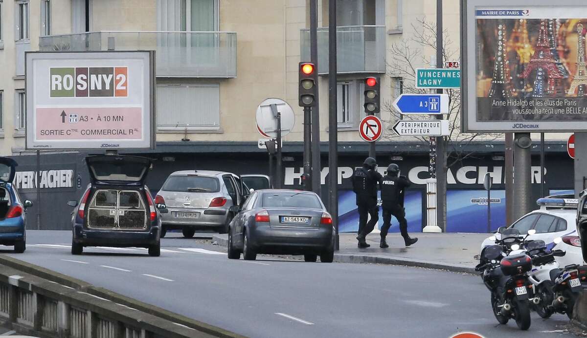 Police officers walk in front of an hostage-taking situation at a kosher market, seen in blue in background, in Paris, Friday Jan. 9, 2015. A police official says the man who has taken at least five people hostage in a kosher market in Paris appears to be linked to the newsroom massacre earlier this week that left 12 people dead. (AP Photo/Francois Mori)