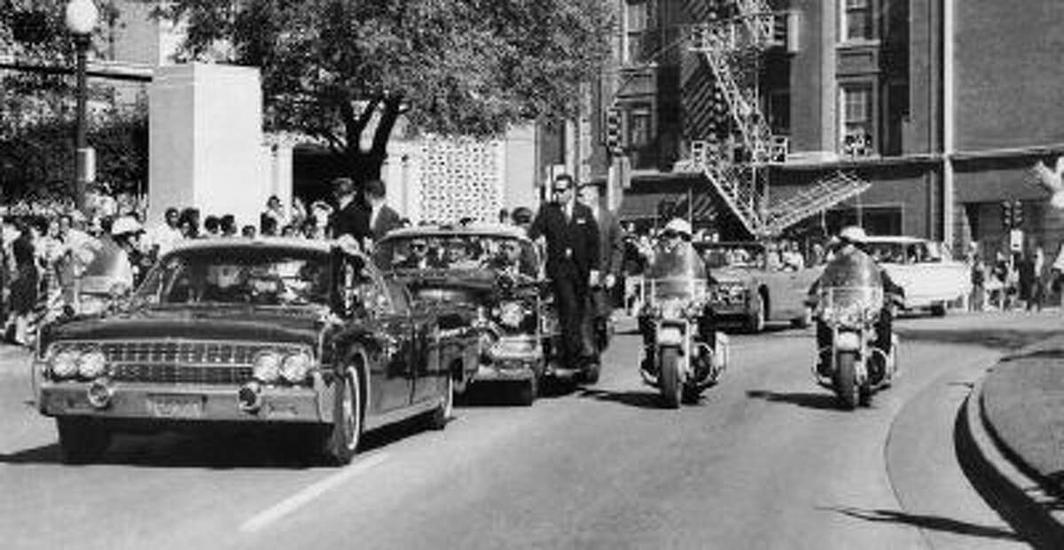 In this Friday, Nov. 22, 1963 file photo, seen through the foreground convertible's windshield, President John F. Kennedy's hand reaches toward his head within seconds of being fatally shot as first lady Jacqueline Kennedy holds his forearm as the motorcade proceeds along Elm Street past the Texas School Book Depository in Dallas.