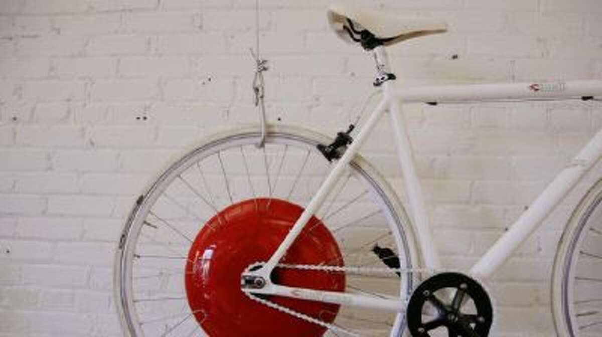 In this Thursday, Dec. 12, 2013 photo, a bicycle fitted with a prototype of the Copenhagen Wheel, the red disk, a human/electric hybrid bicycle engine, hangs on a wall at Superpedestrian, the hybrid engine's manufacturer in Cambridge, Mass.