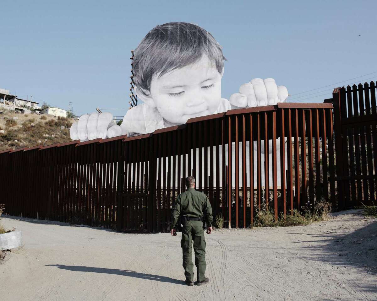 A border patrol officer on the U.S. side of the Mexico border wall, across from an art installation by artist JR, based on his photo of a 1-year-old visible from the California side near Tecate, Mexico. The art might be a metaphor for the future of the American workforce, which will be dependent on immigrants and their descendants.