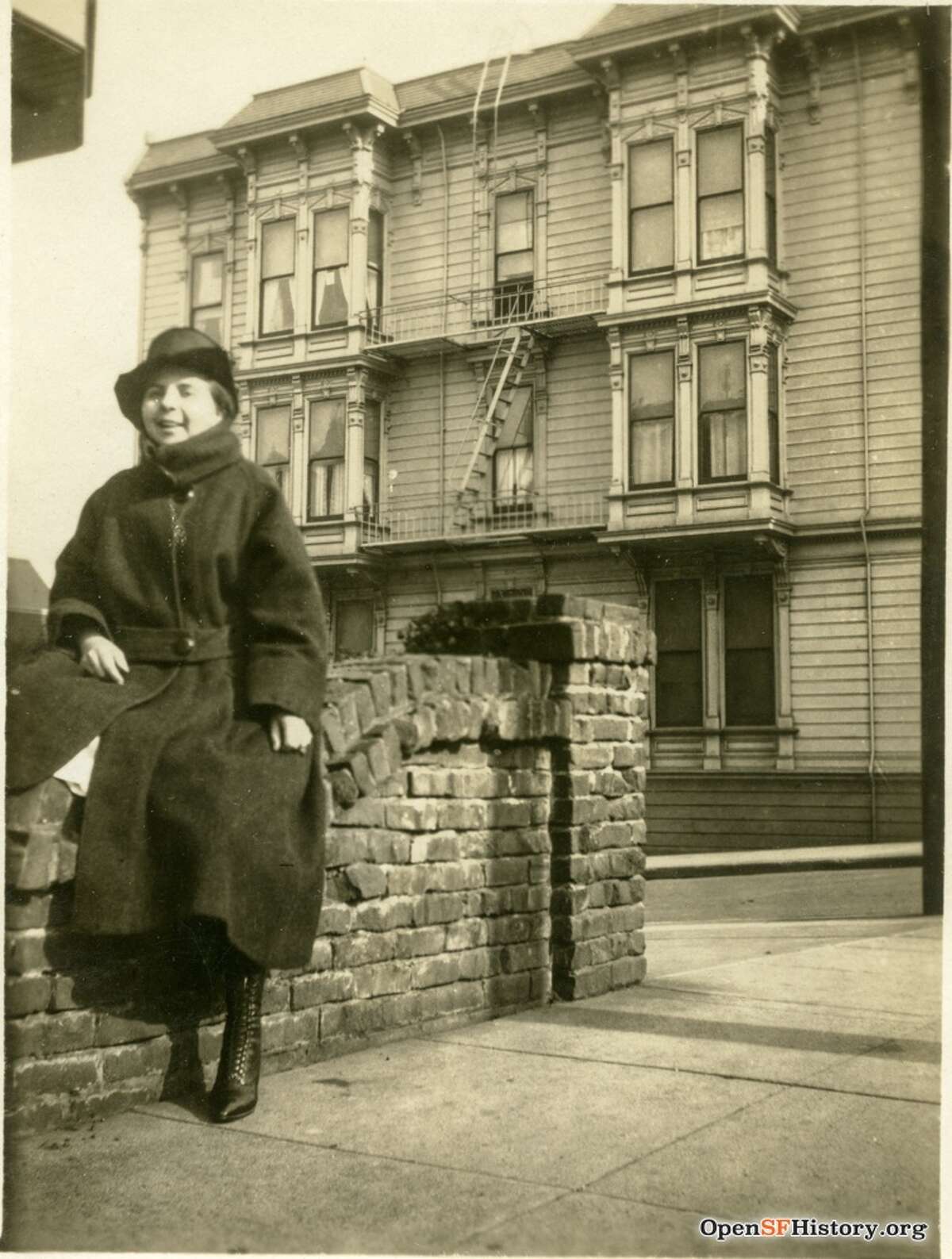 A woman posing against brick wall, with 1192 Haight in the background. Photo courtesy of OpenSFHistory.