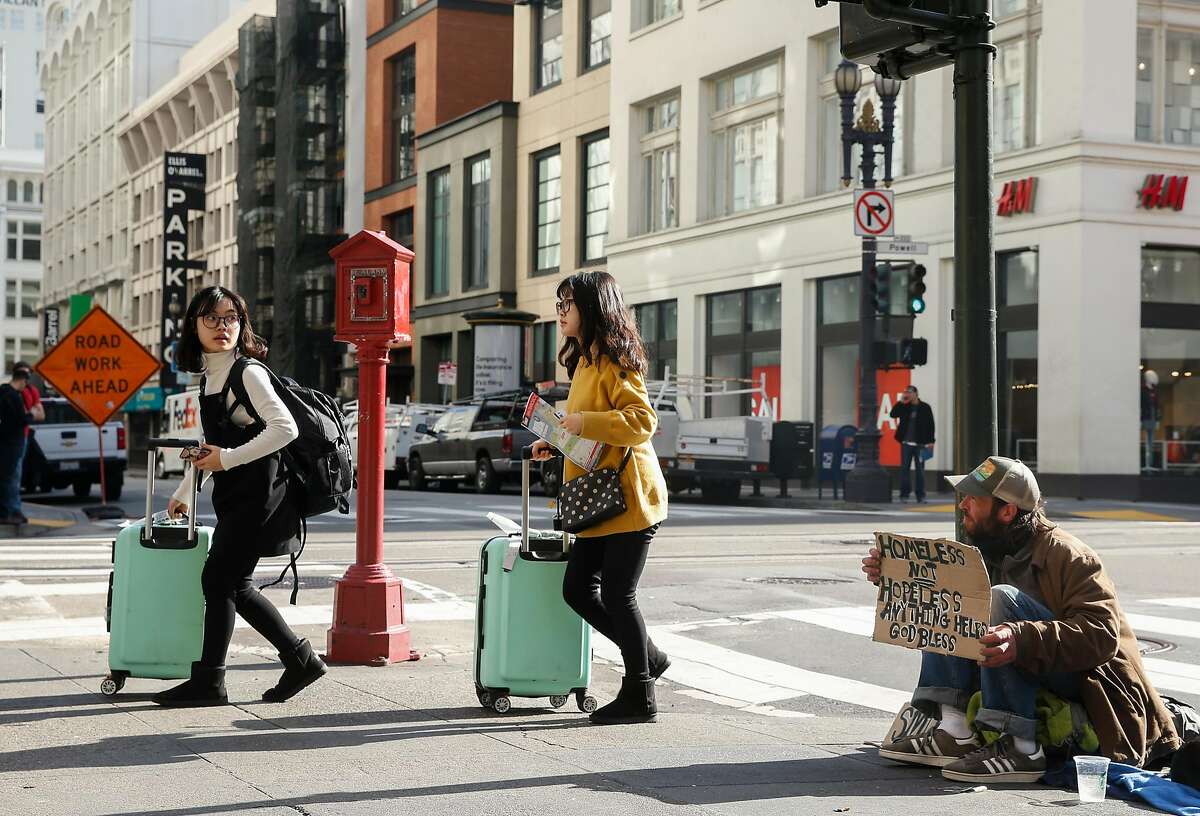 Two women with suitcases walk past Abram Lange, a homeless man originally from Eugene, Oregon, on the corner of Powell and O'Farrell streets Tuesday, Jan. 23, 2018 in San Francisco, Calif.