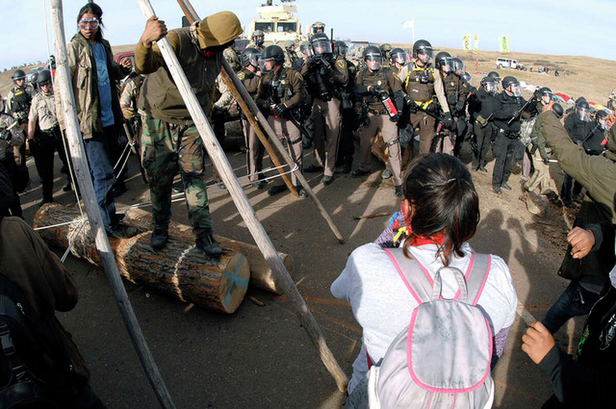 Mike McCleary | The Bismarck Tribune (AP) Protesters shield their faces as a line of law enforcement officers holding large canisters with pepper spray shout orders to move back during a standoff in Morton County, North Dakota. On the same day seven defendants celebrated acquittal in Portland, Oregon, for their armed takeover of a federal wildlife refuge, nearly 150 protesters camped out in North Dakota to protest an oil pipeline were arrested.