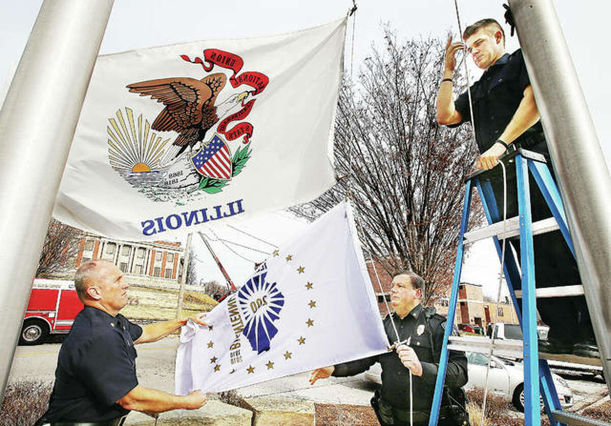Alton Fire Chief Bernie Sebold, left, and Alton Police Deputy Chief Terry Buhs, right, unfold the new Illinois Bicentennial Flag as probationary firefighter Richard Sherman, far right, prepares to raise it Monday underneath the regular Illinois flag at Alton City Hall. The flag ceremony, like several across the area, was a brief part of a noon event to mark the kickoff a year-long celebration of the state’s bicentennial, which occurs in December 2018.