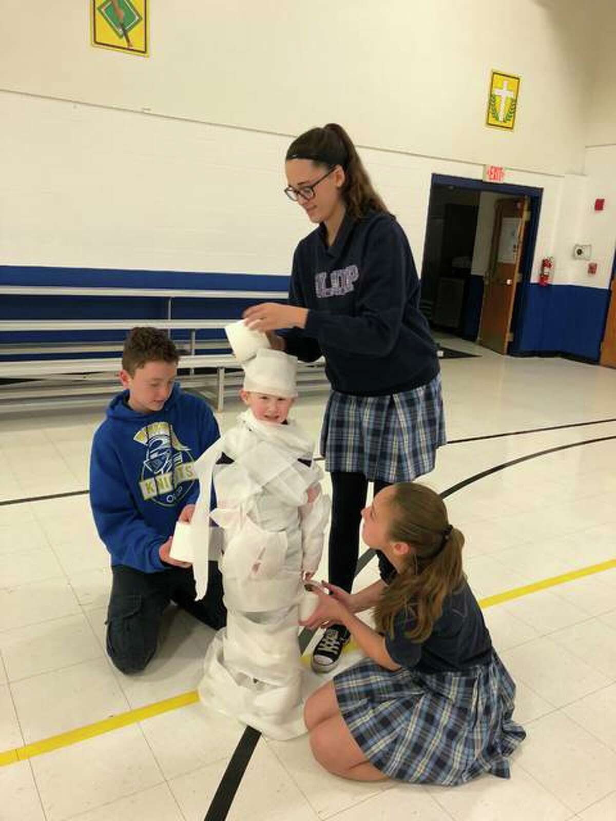 Students, using toilet paper, wrap-up a kindergartner during a recent Christmas party, to make him look like a snowman. The children, students at Our Lady Queen of Peace School, are members of the Buddy Program, which pairs an eighth-grader to help a kindergartner for the year.