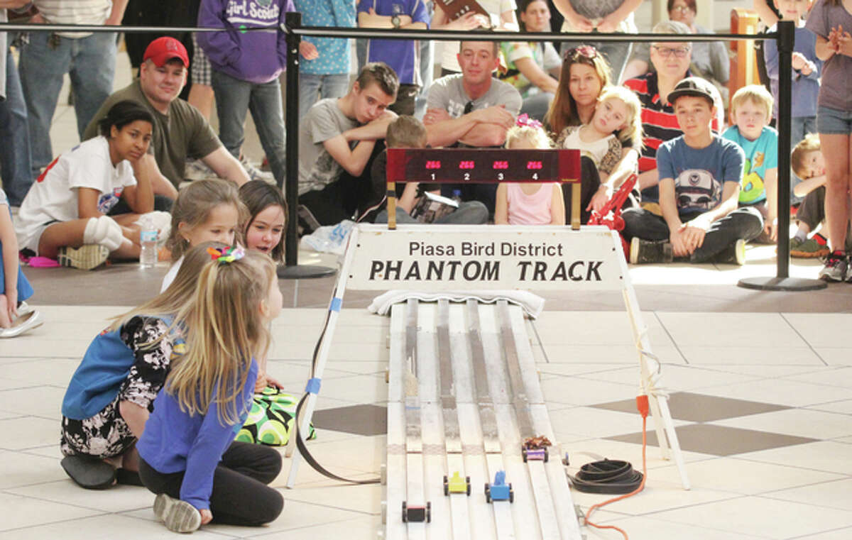 Competitors sitting next to the track and the crowd behind the barrier watch as girls compete in pinewood derby racing Saturday at Alton Square.