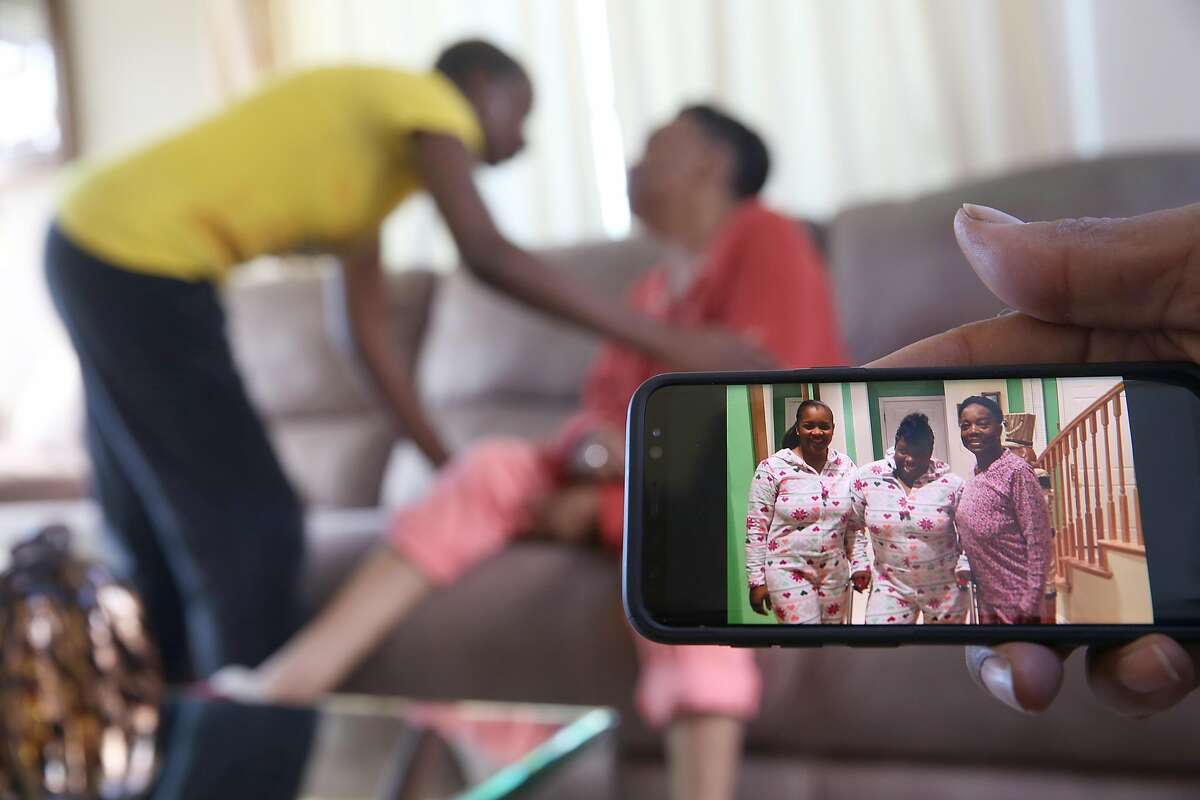 Home health care provider Sonia Wright (left in background) says goodbye to Margie Cherry (middle in background) as her husband Johnny Cherry (right) shows pictures of Margie (wearing pink) with their two grown daughters at left on phone at home on Friday, February 9, 2018, in San Francisco, Ca.