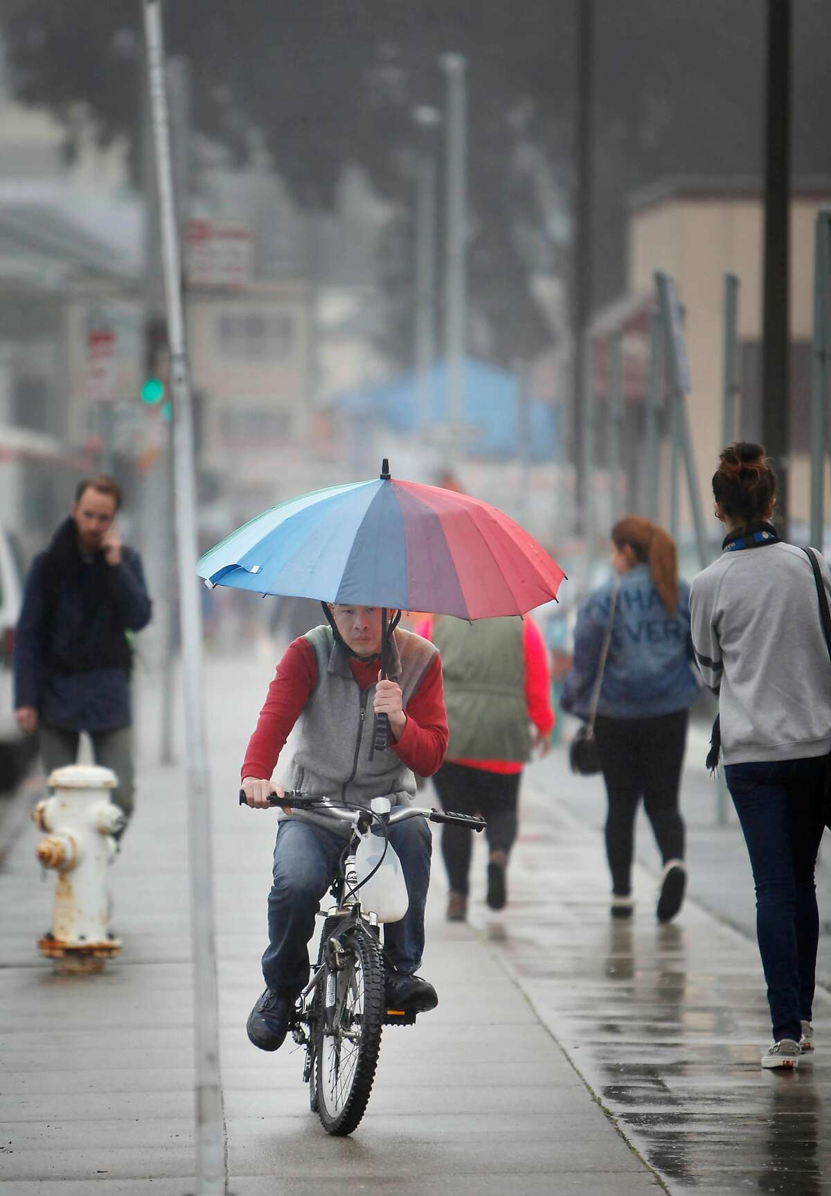 A bicyclist carries an umbrella in a light rain as he rides on the sidewalk along Phelan Avenue on Wednesday, January 29, 2014 in San Francisco, Calif.