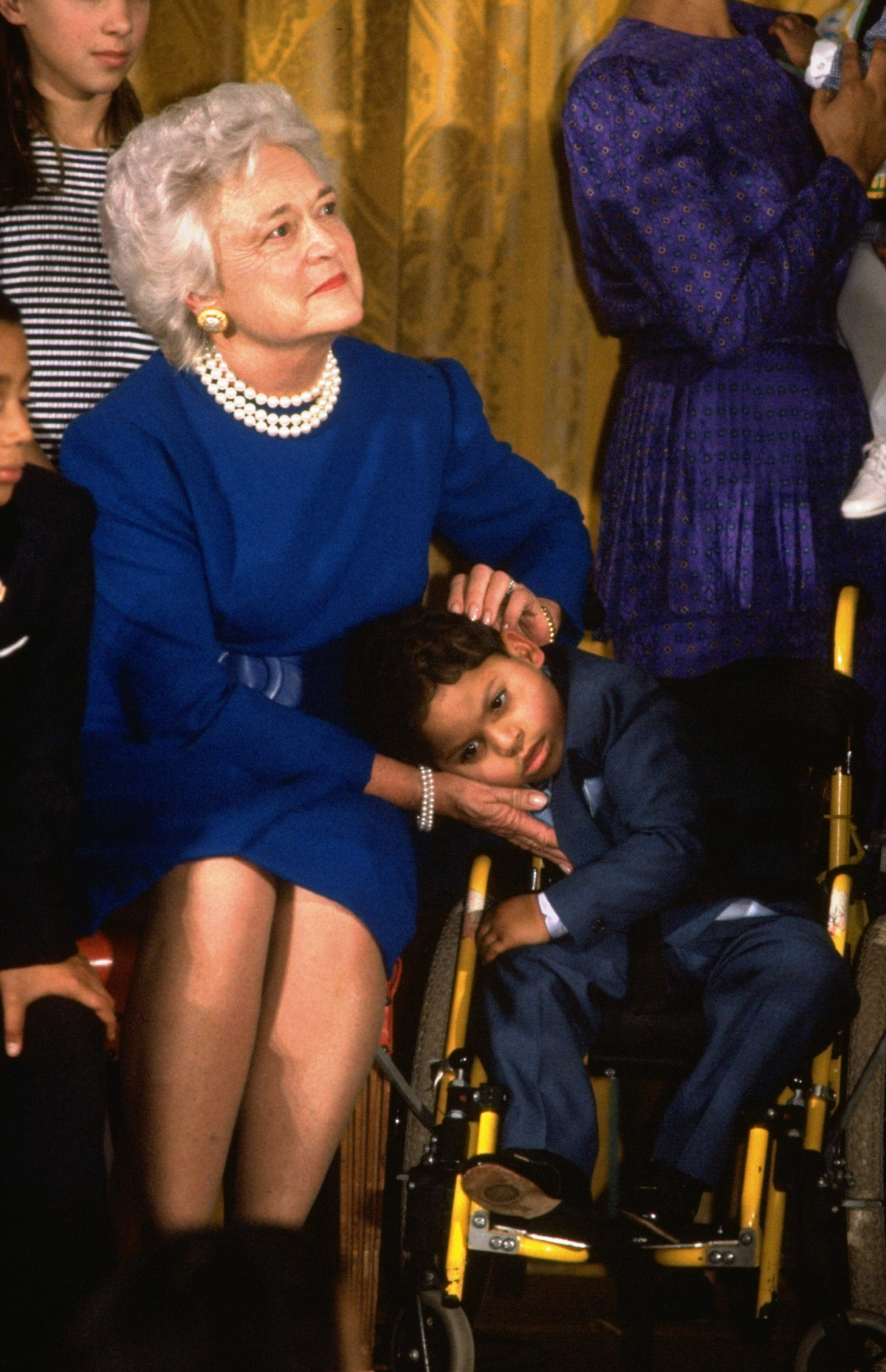 Barbara Bush holding Angel Orsi with affection as the little wheelchair-bound boy puts his head in her lap during a White House Celebration of Children.