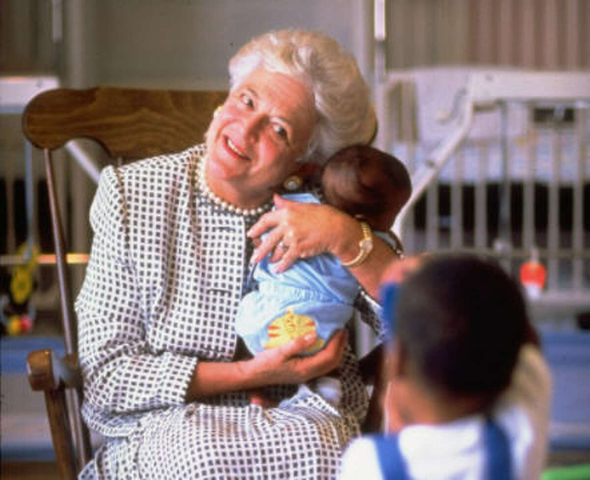 First Lady Barbara Bush holding a baby while two-year-old child takes photo with a toy camera at a hospice for children with AIDS. See more photos of the first lady through the years...