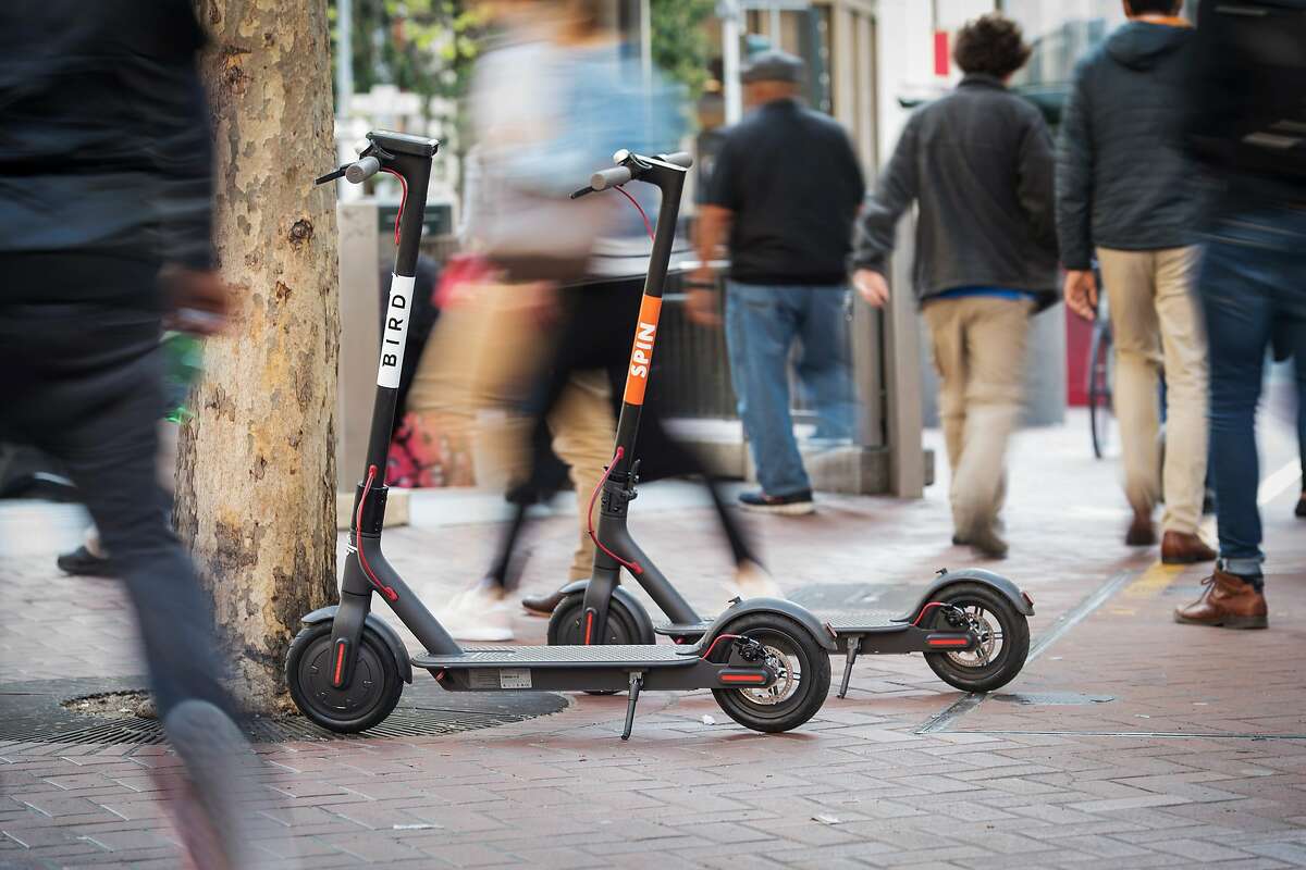 Pedestrians walk past a Bird Rides Inc., left, and Skinny Labs Inc. SpinBikes shared electric scooters parked on Market Street in San Francisco, California, U.S., on Friday, April 13, 2018. GPS-enabled scooters and bicycles are spreading across several major U.S. cities, driven by a wave of venture capital into a handful of companies. Policymakers are scrambling to find ways to regulate the great scooter boom of 2018. Photographer: David Paul Morris/Bloomberg