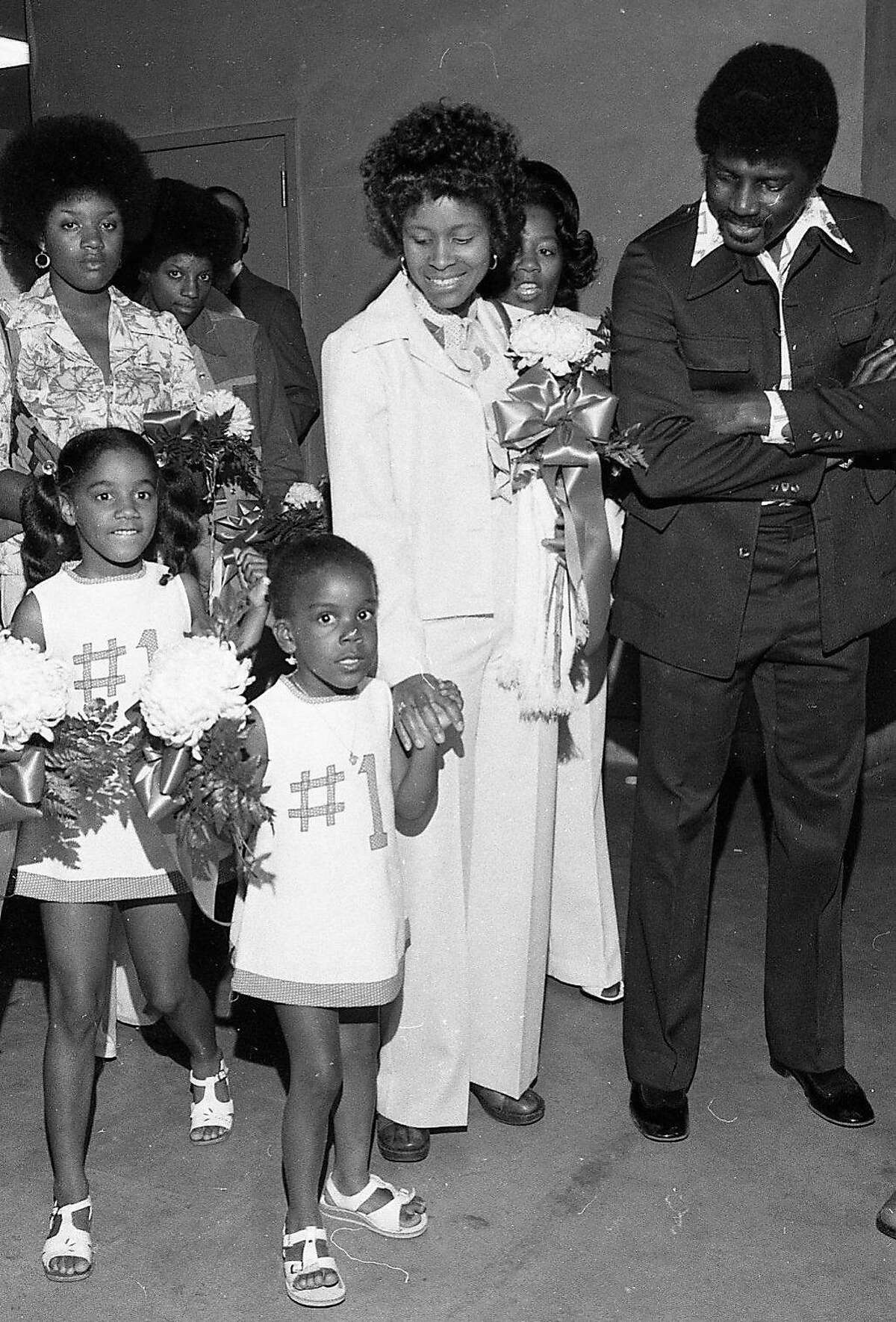 1975年5月29日，在奥克兰体育馆举行的金州勇士队颁奖典礼上，赢得NBA总冠军后，助理教练乔·罗伯茨的女儿埃里卡·阿特尔斯和乔迪·罗伯茨身着白色礼服配蓝色#1