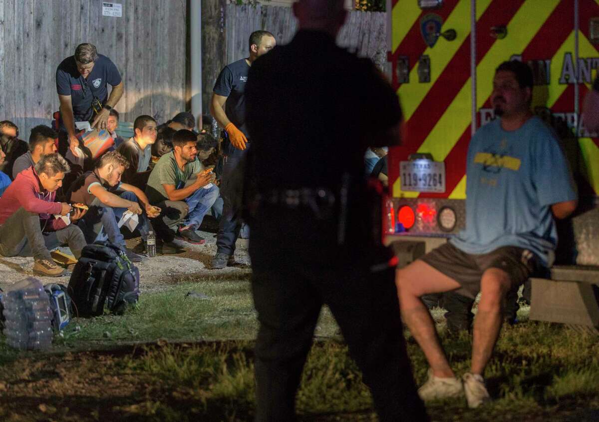 A man in handcuffs is watched by a police officer as apparently undocumented immigrants sit on the ground Tuesday night, June 12, 2018 after being found in the back of an 18-wheel truck near loop 410 and Broadway.
