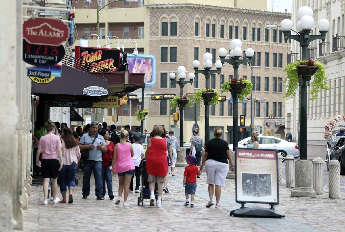 People walk on the Alamo Street sidewalk past Alamo Plaza tourist attractions.