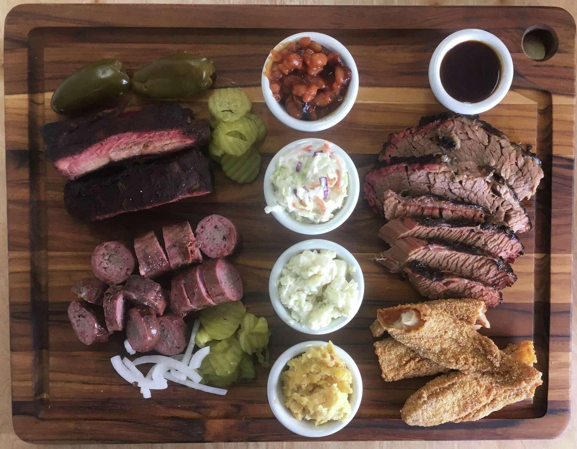 A wooden board laden with various BBQ meats including ribs, brisket, and sausage, along with sides like coleslaw, pickles, and baked beans.
