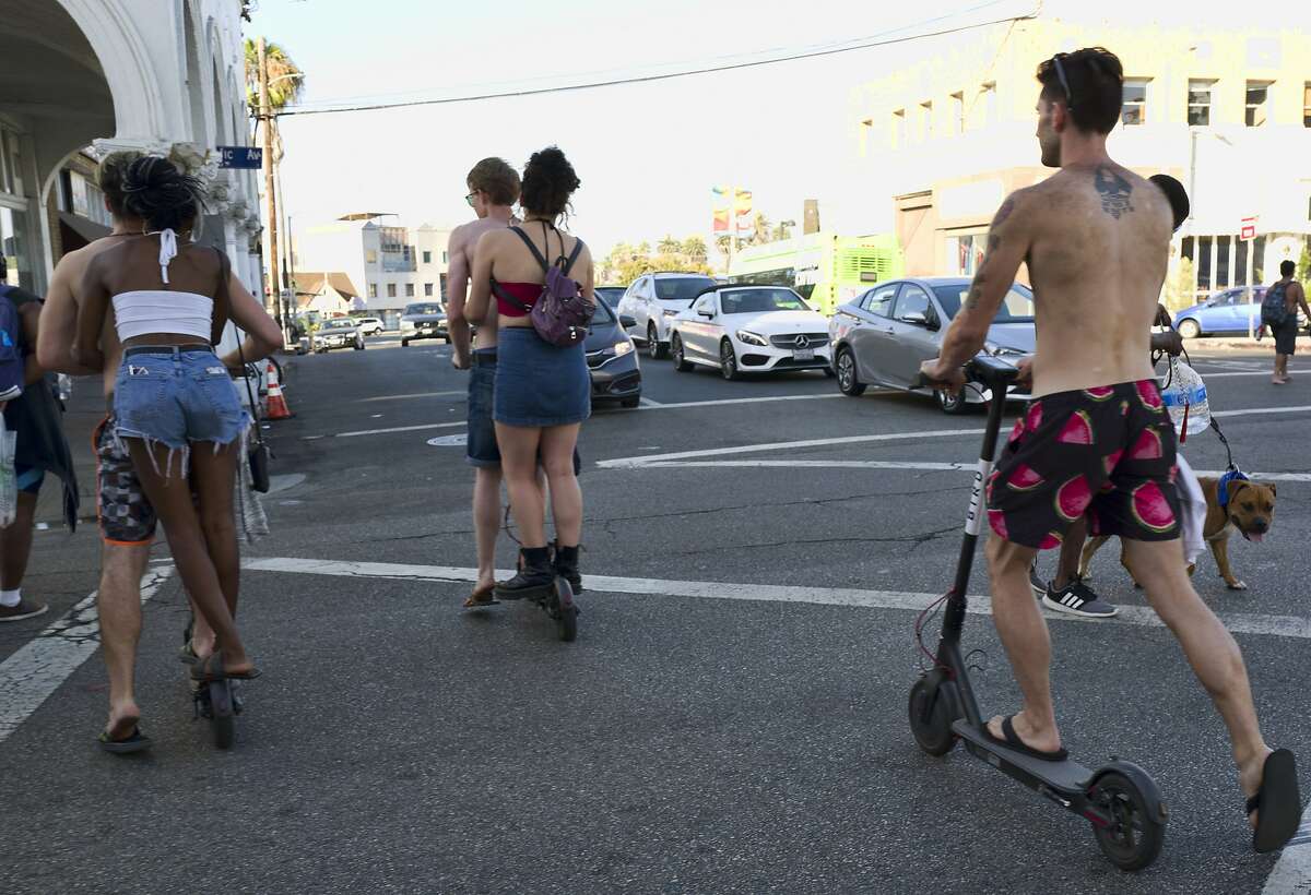 FILE - In this July 24, 2018, file photo, riders make their way across a street on Bird electric scooters in the Venice Beach section of Los Angeles. Californians could ride motorized scooters without helmets under legislation headed to Gov. Jerry Brown. Lawmakers in the California Assembly passed the bill Wednesday, Aug. 29, 2018. It would let adults ride the devices without helmets and allow riders to travel on faster-moving streets. (AP Photo/Richard Vogel, File)
