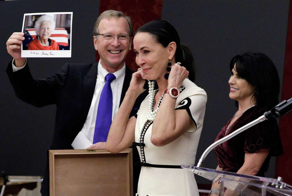 Neil Bush holds a photo of his mother, former first lady Barbara Bush wearing the same pearl necklace that Sue Smith models that was given to Sue by her husband as Maria Bush looks on at the Barbara Bush Literacy Foundation's "Celebration of Reading" party at the home of Sue and Lester Smith in Houston, TX, Dec. 7, 2017.