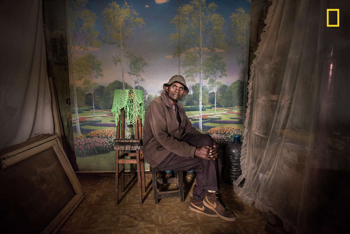 Winners of the 2018 National Geographic Photo Contest, 1st Place, People I was looking to do a series of portraits showing people wearing their Sunday best when I made this photo of David Muyochokera. It was taken on his last Sunday working as a photographer at Weekend Studio, in Kibera—a large shantytown in Nairobi, Kenya. Myfriend Peter, a local resident, had pointed me to the photo studio just as I was about to leave the area. It was a stunning space, with whimsical backdrops and natural light coming through the doorway. David had worked there for 37 years, but Weekend Studio was about to close permanently. Phone cameras were so common now, he said, and fewer people wanted studio portraits. David planned to retire and return home to the countryside. I  was troubled by the studio's imminent closure, so I eventually took over the rent. A portrait of David now hangs at Weekend Studio in his memory. Link: https://yourshot.nationalgeographic.com/photos/12670253