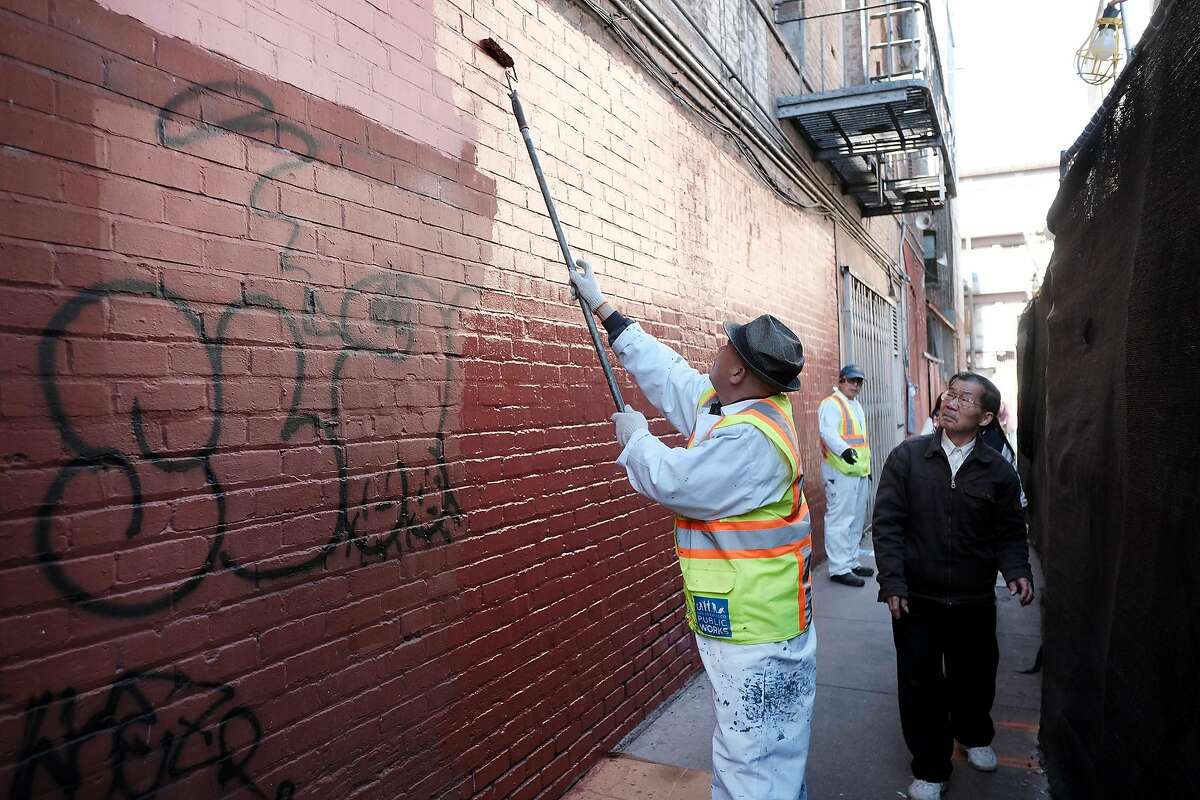 People pass by as Department of Public Works graffiti unit employee David Yu paints over graffiti in Hang Ah Alley in San Francisco, Calif., on Thursday, December 27, 2018.