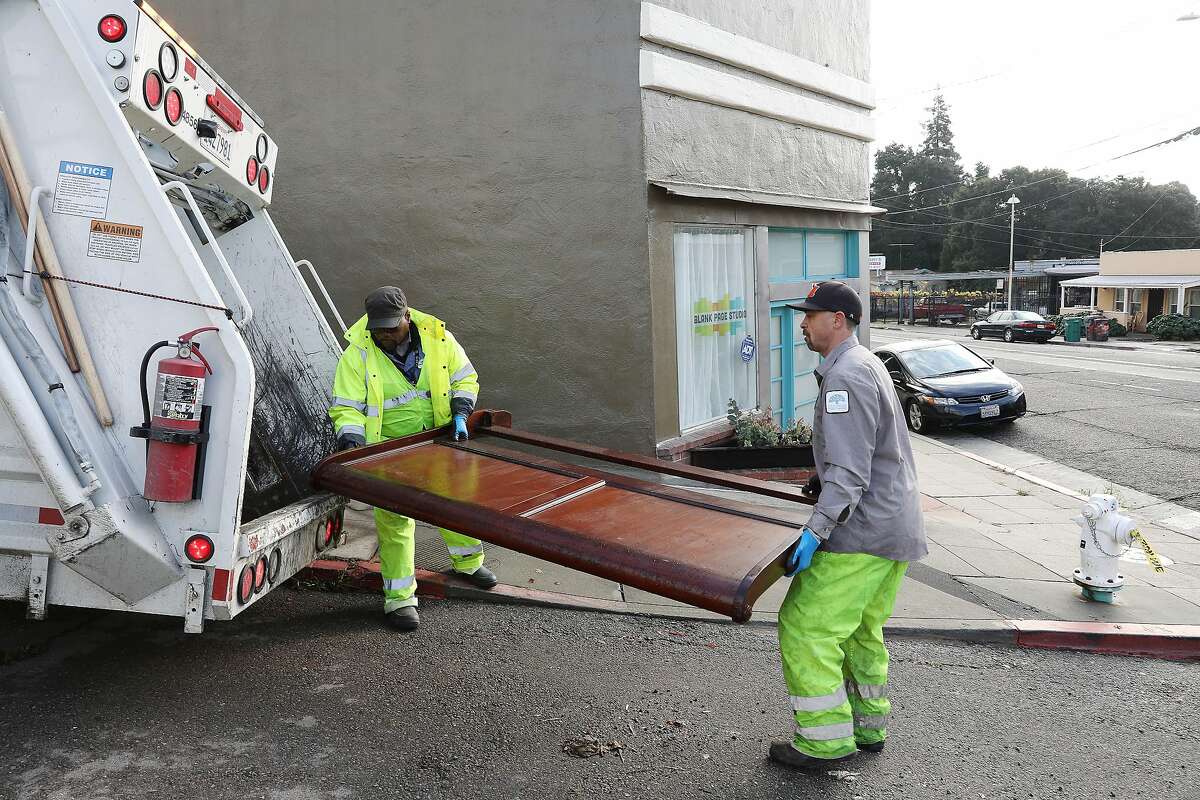 Ayinde Osayaba (l to r) and David Conti, both Keep Oakland Clean and Beautiful public maintenance worker, carry a headboard to the back of a garbage truck on Monday, December 17, 2018 in Oakland, Calif.